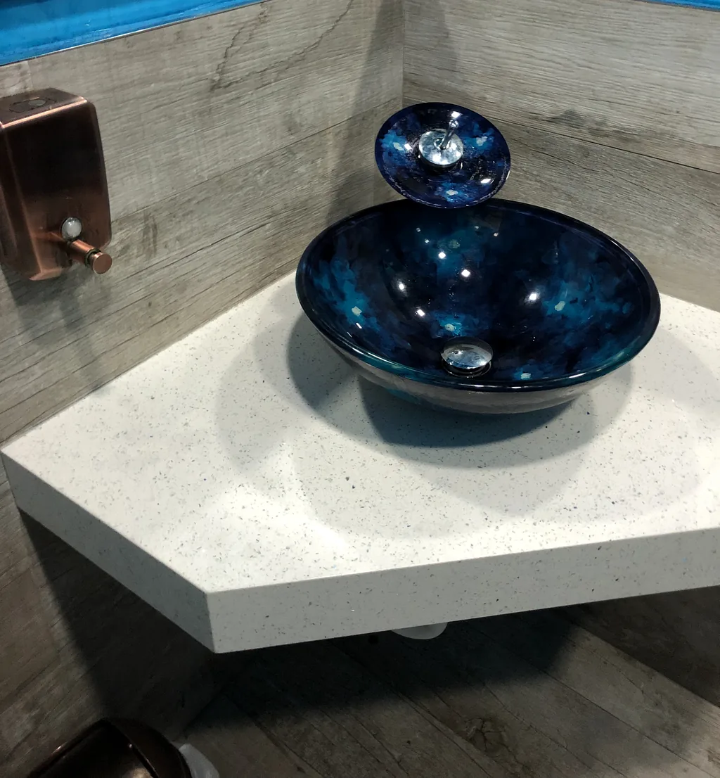 Blue glass vessel sink on a white quartz corner counter against wood-look tile